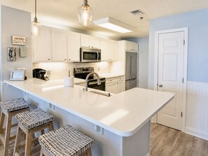 A kitchen with white cabinets, a stainless steel refrigerator, a microwave, and a large island with stools. - 204 Palacio, low floor professionally decorated gulf front condo! (Pensacola)