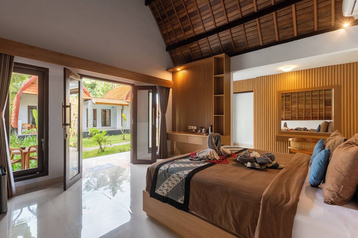 A spacious bedroom with a large bed, a wooden wardrobe, and a balcony with a view of a house and greenery.