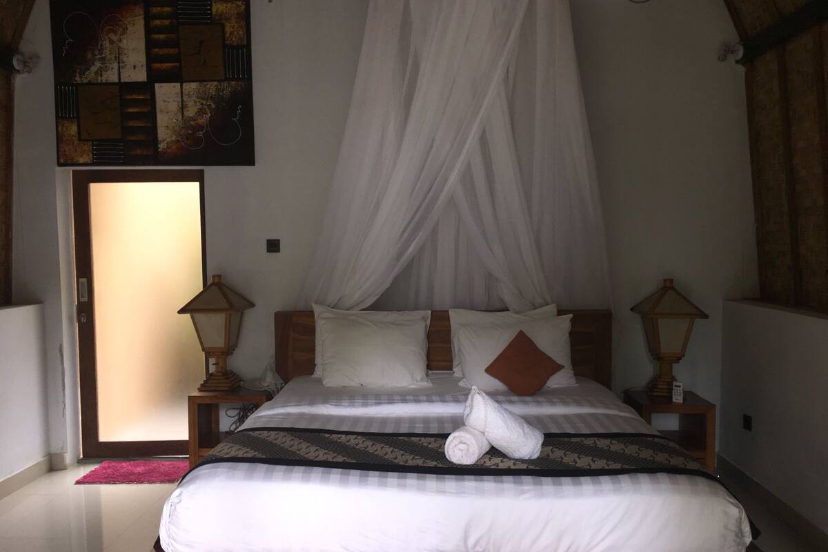 A bedroom with a canopy bed, two bedside lamps, a wooden headboard, and a view of the outside.