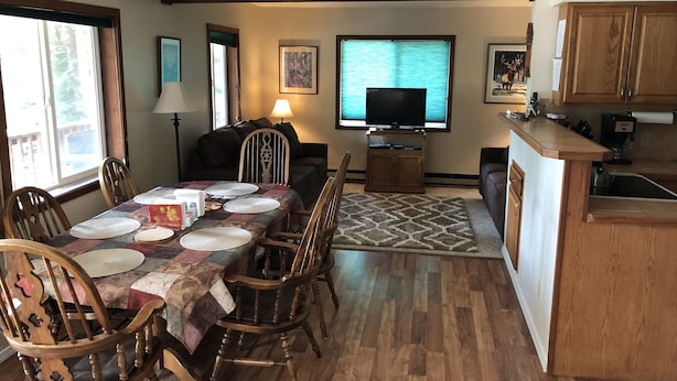 A dining area with a wooden table set for four, surrounded by wooden chairs. A television is mounted on the wall above the table. - Ketchikan Northern Retreat - 3 Bedroom Apartment on Large Private Lot (Ketchikan)