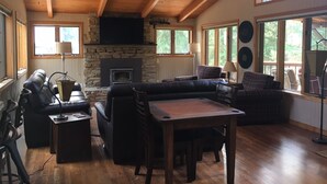 A cozy living room with a stone fireplace, wooden ceiling beams, and large windows offering a view of the outdoors.