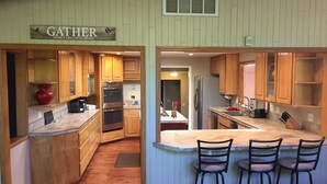A kitchen with wooden cabinets, a central island with bar stools, a red vase, and a sign that reads "GATHER WITH A GRATEFUL HEART".