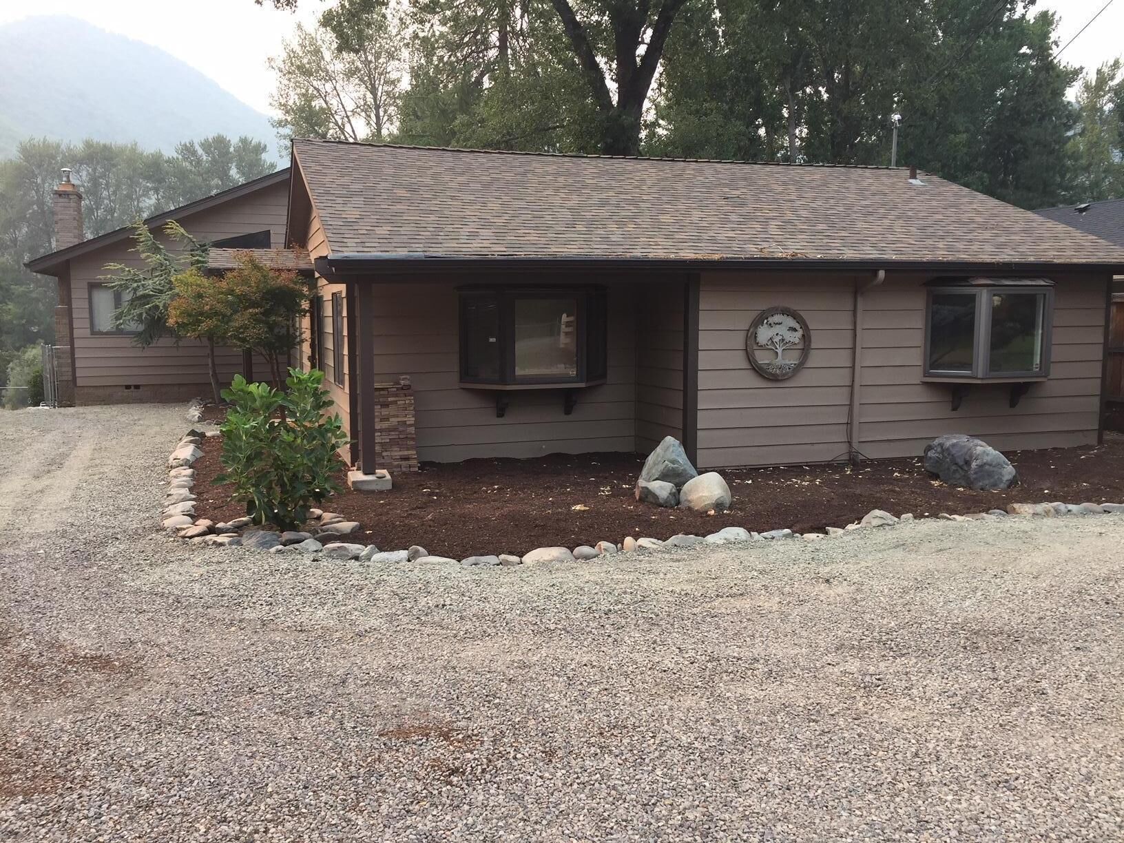 A single-story house with a gravel driveway, a small garden with rocks, and a mountain in the background.