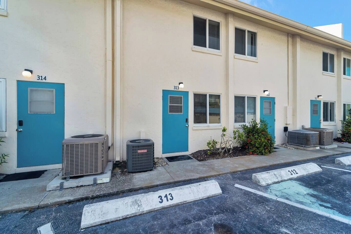 A row of motel rooms with numbered doors, air conditioning units, and a parking lot with marked spaces.