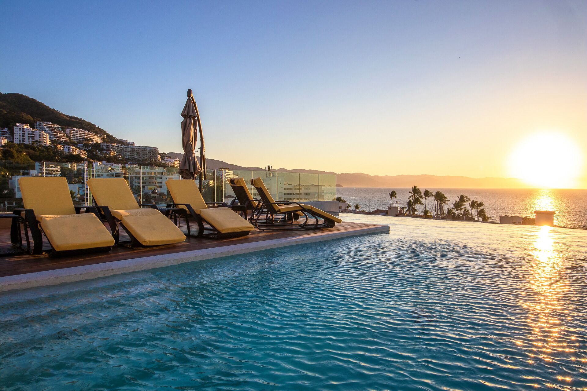 Zona junto a la piscina con sillones de descanso y barandilla de cristal con vista al atardecer sobre el mar.
