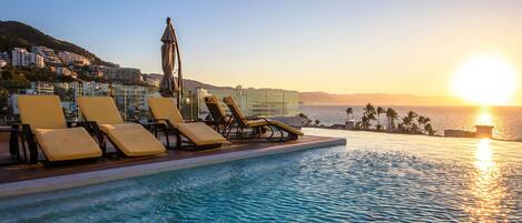 Zona junto a la piscina con sillones de descanso y barandilla de cristal con vista al atardecer sobre el mar.