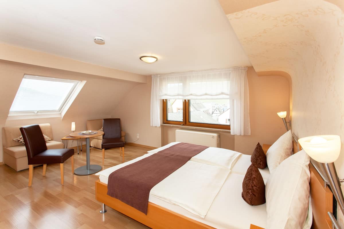 A modern hotel room with a large bed, a sitting area with a chair and table, a window with curtains, and a radiator.