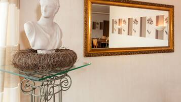 A bust sculpture on a glass table with a woven nest beside it, a gold-framed mirror on the wall, and a room with floral wallpaper and a dining area in the reflection.