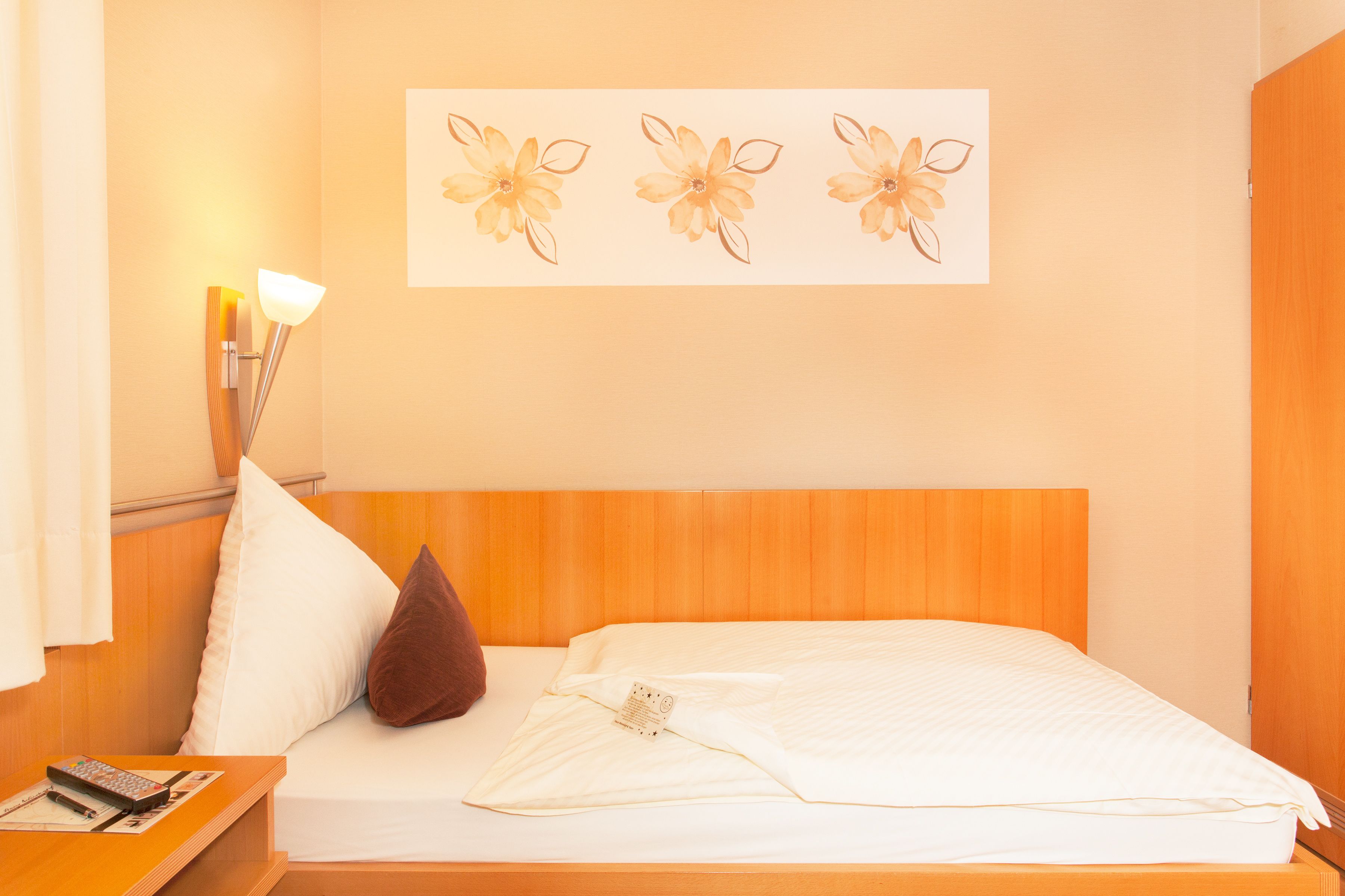 A hotel room with a wooden headboard, a bed with white bedding, a brown pillow, a nightstand with a remote control, and a wall-mounted lamp.