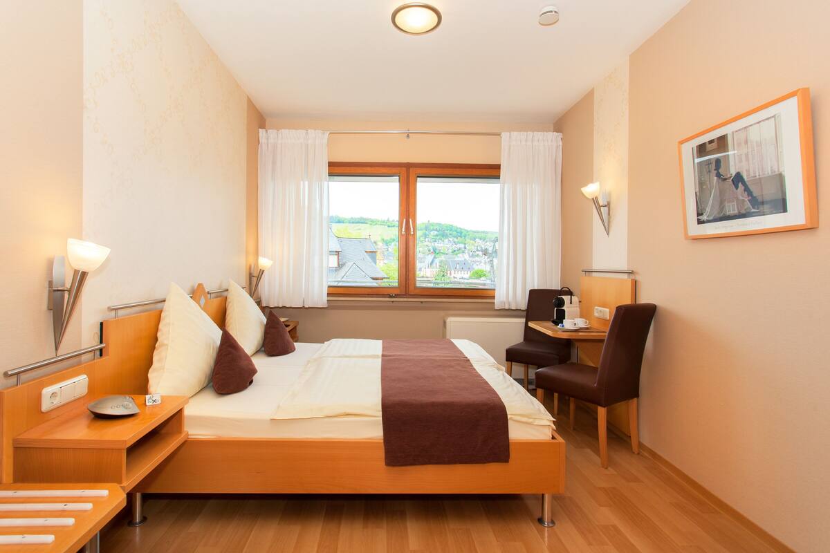 A hotel room with a bed, bedside table, chair, desk, and a view of the outside.