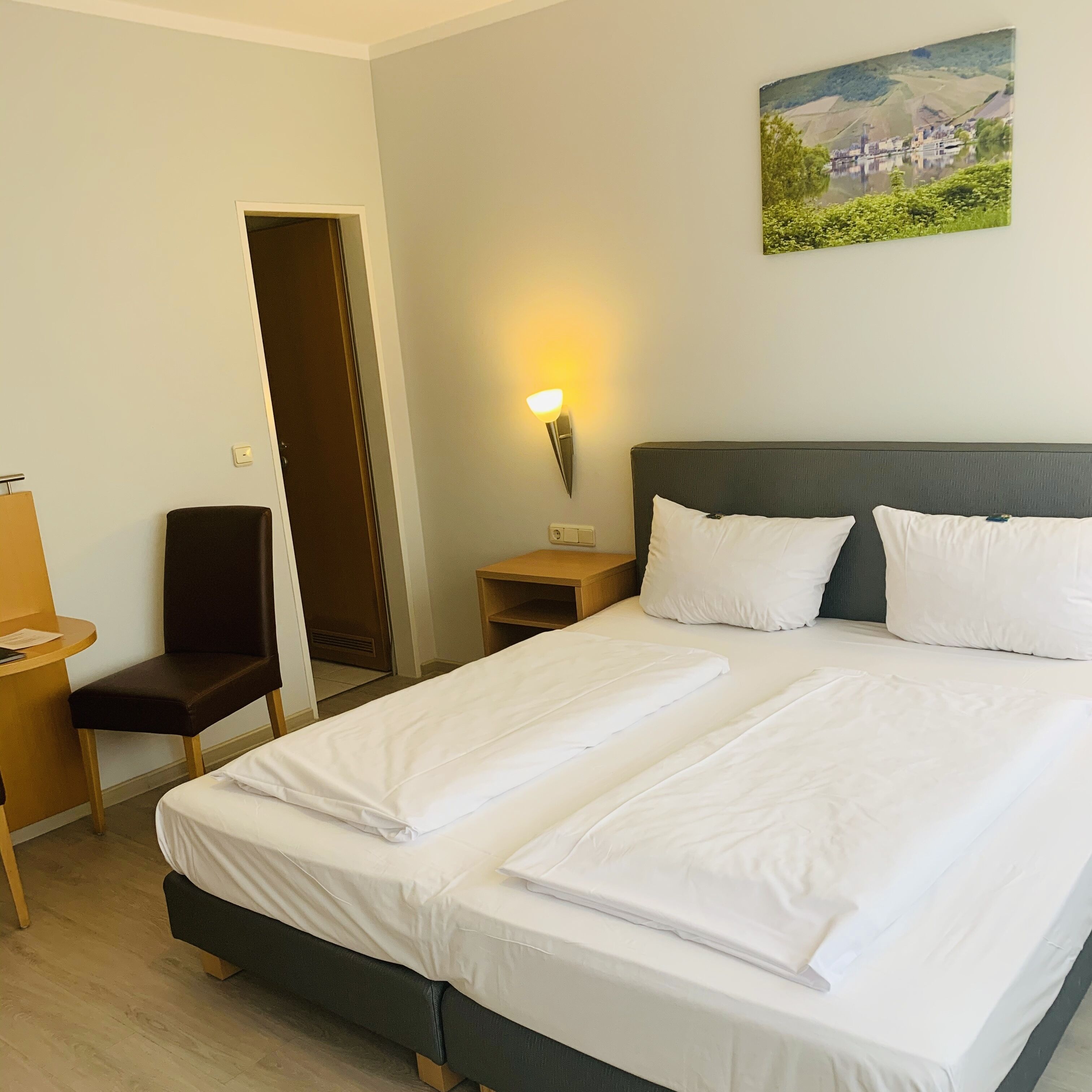 A hotel room with a bed, a chair, a desk, and a painting on the wall.