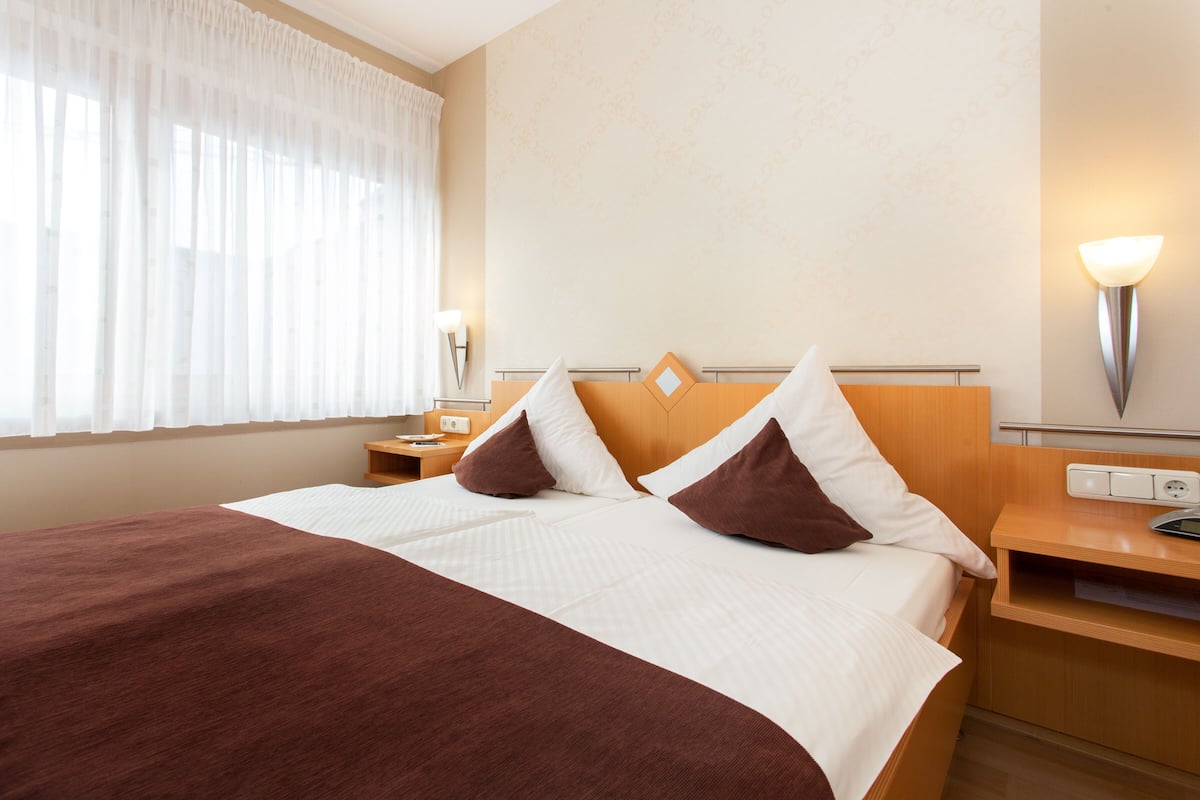A hotel room with a large bed, two pillows, a nightstand, a lamp, and a wall-mounted light.