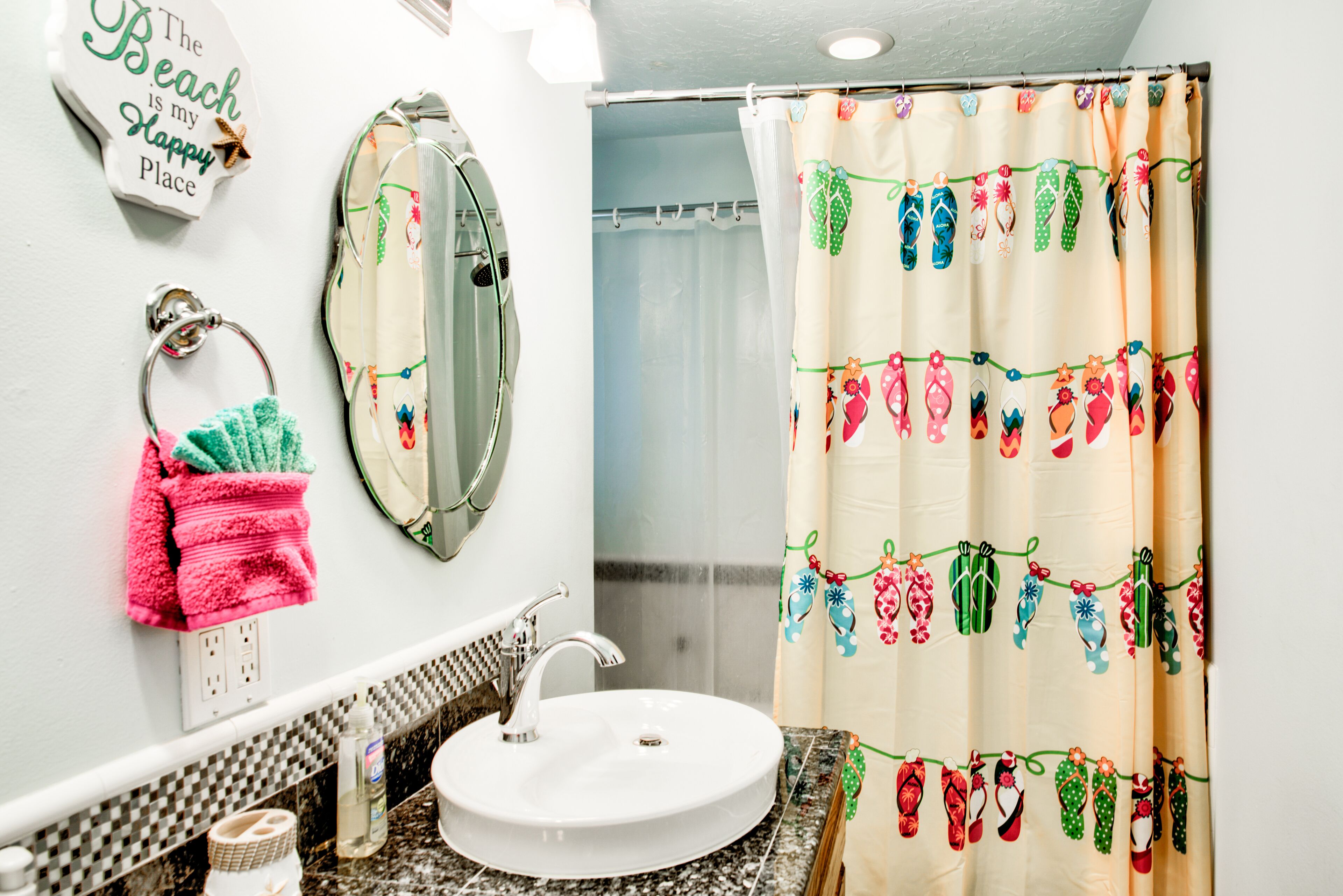 Une salle de bain avec un lavabo blanc, un miroir et un rideau de douche orné d’un motif sur le thème de la plage.
