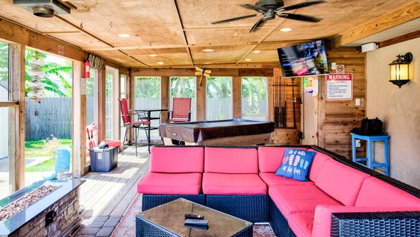 A covered outdoor area with a pool table, a red sofa, and a television mounted on the wall.