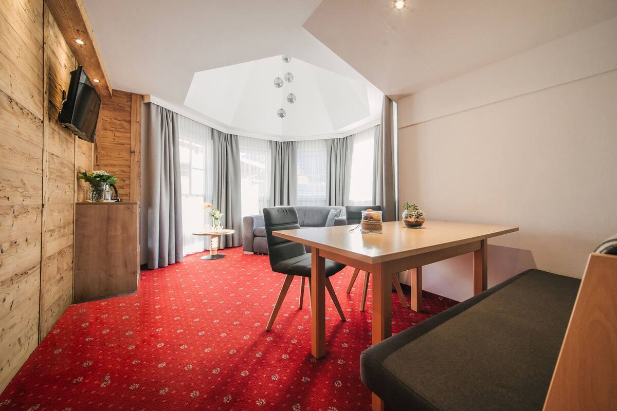 A room with a wooden wall, a red carpet, a dining table with chairs, a sofa, and a TV mounted on the wall.