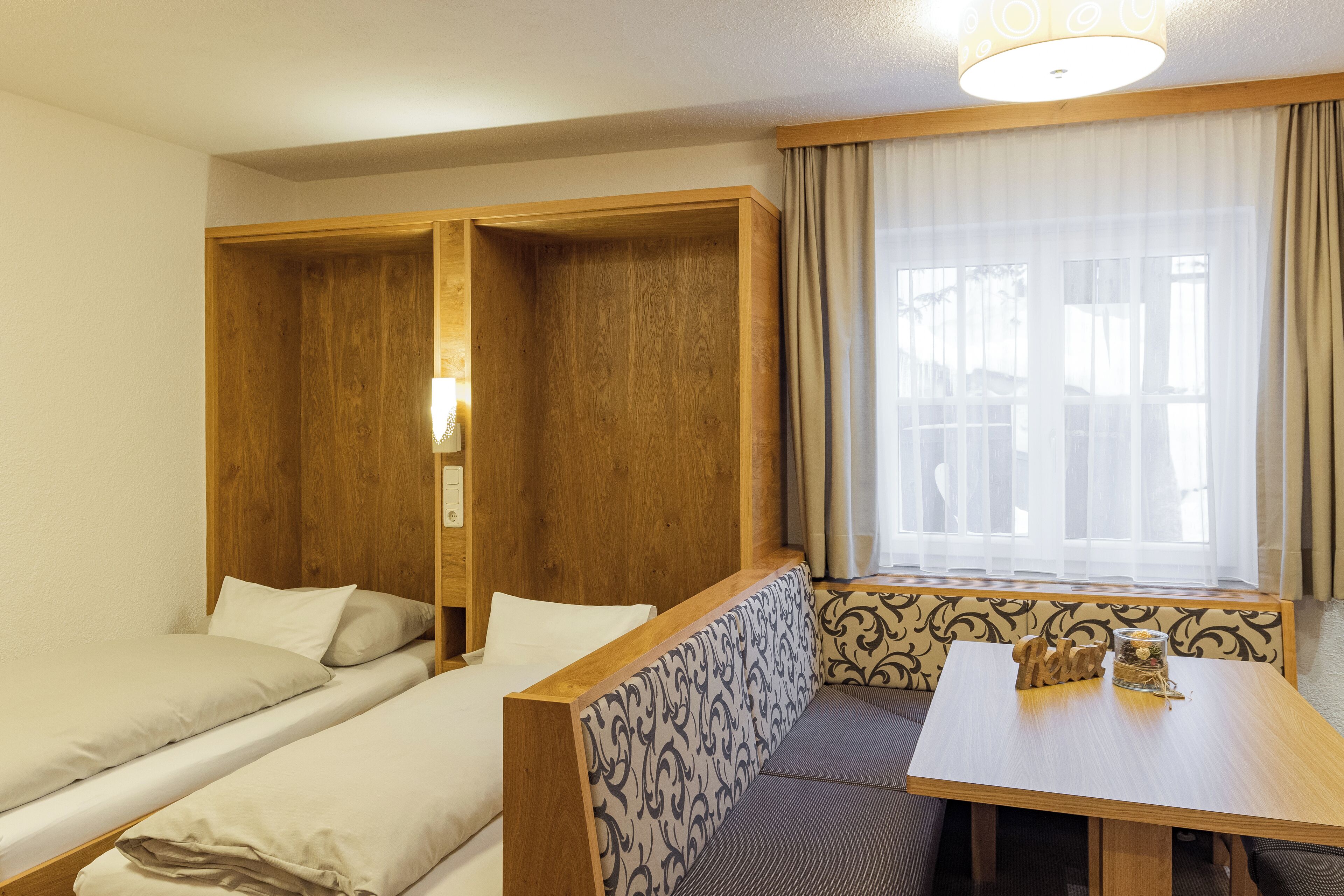 Quarto com duas camas, cabeceira de madeira, mesa com um banco e janela com cortinas.