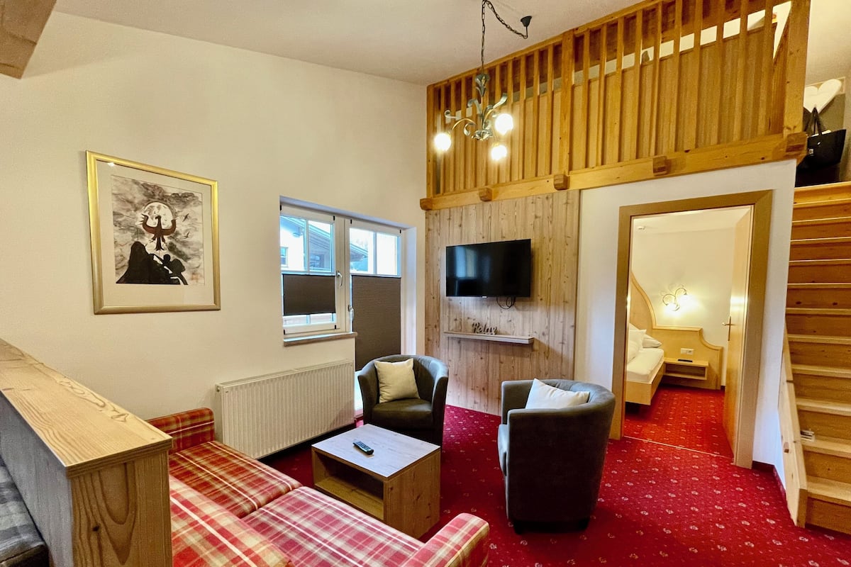 A cozy living room with a red carpet, a sofa, two armchairs, a television, and a wooden ceiling.