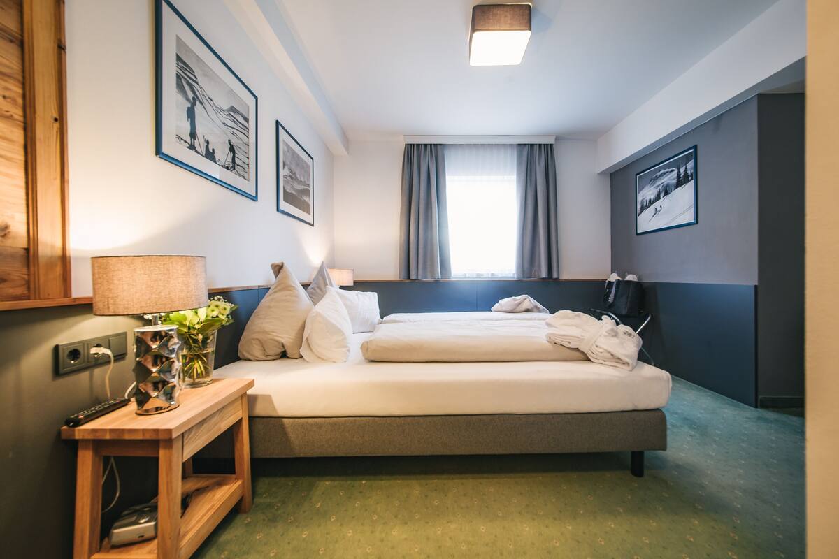 A hotel room with a bed, a bedside table with a lamp, a vase with flowers, and framed pictures on the wall.