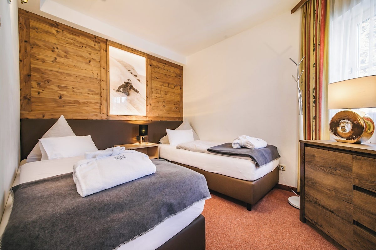 A hotel room with two beds, wooden headboards, a large framed picture, a bedside table with a lamp, and a dresser with a decorative item.