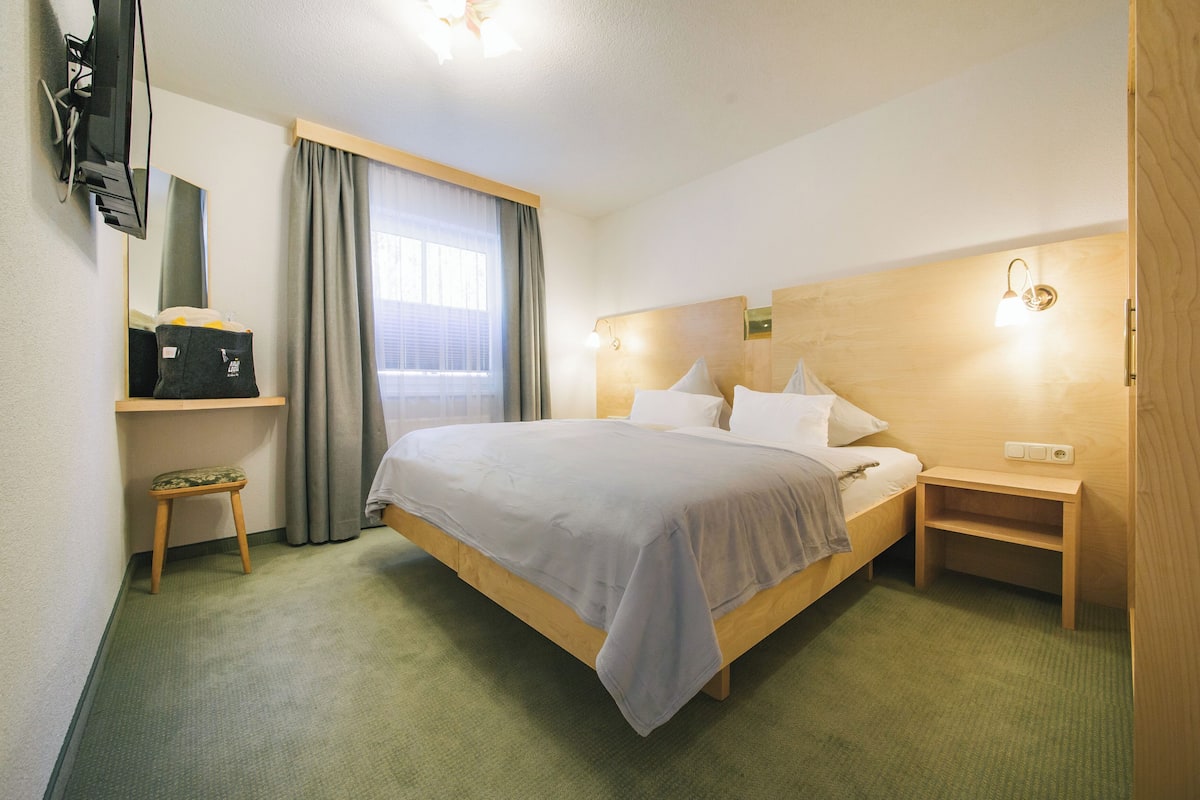 A hotel room with a bed, a TV mounted on the wall, a small table, and a nightstand.