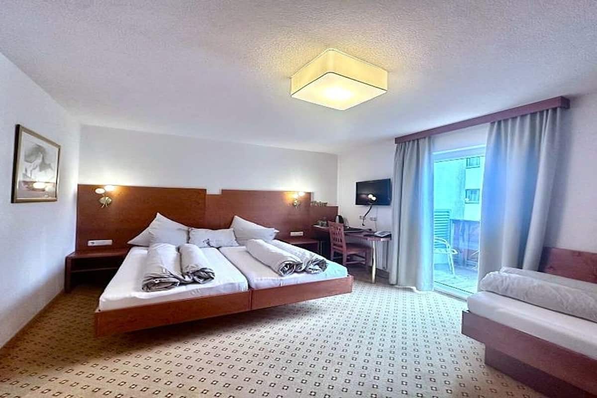 A hotel room with a large bed, a desk, a chair, a TV, and a balcony with curtains.