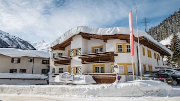 Un immeuble à plusieurs étages, dont le toit et les balcons sont recouverts de neige, entouré de neige et de montagnes.