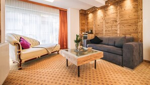 A living room with a beige sofa, a gray sofa, a glass coffee table, and a wooden accent wall. - Arlen Lodge Hotel (Sankt Anton am Arlberg)