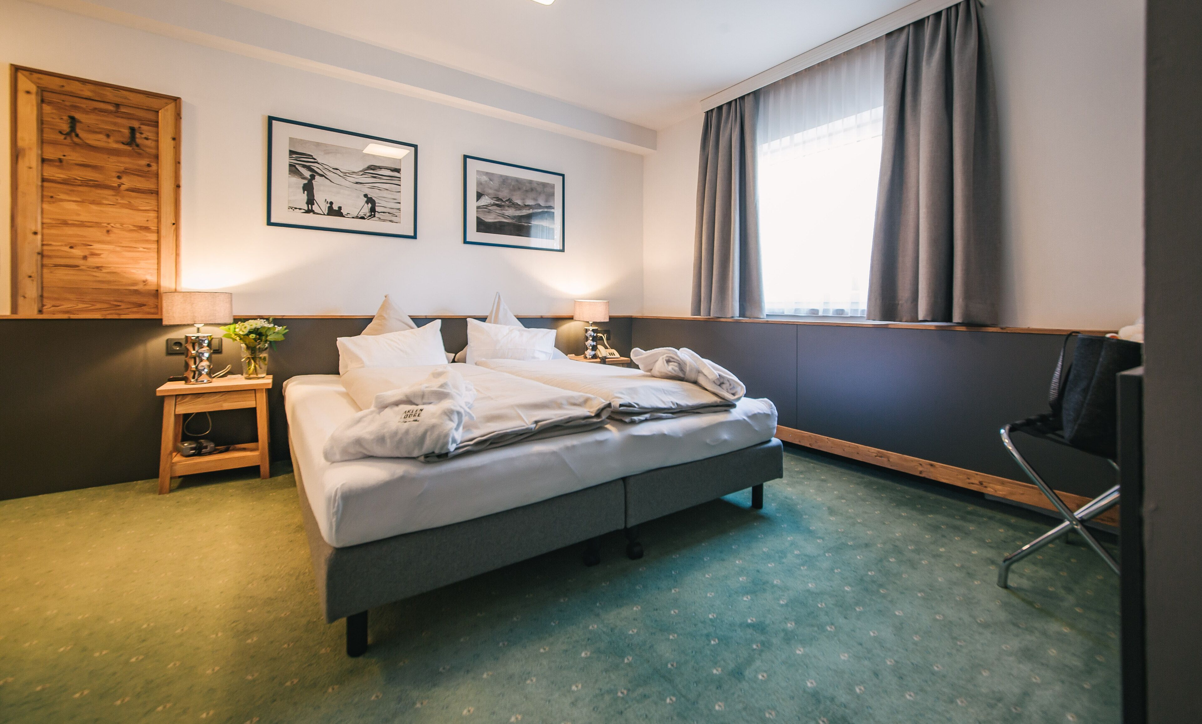 Habitación de hotel con cama, mesita de noche, una silla, ventana con cortinas y dos cuadros en la pared.