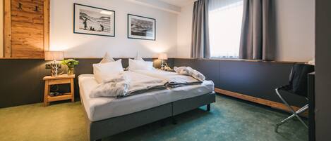 Habitación de hotel con cama, mesita de noche, una silla, ventana con cortinas y dos cuadros en la pared.