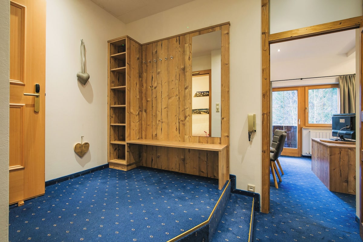 A room with a wooden wardrobe, a mirror, a blue carpet, and a view of the outside through a window.
