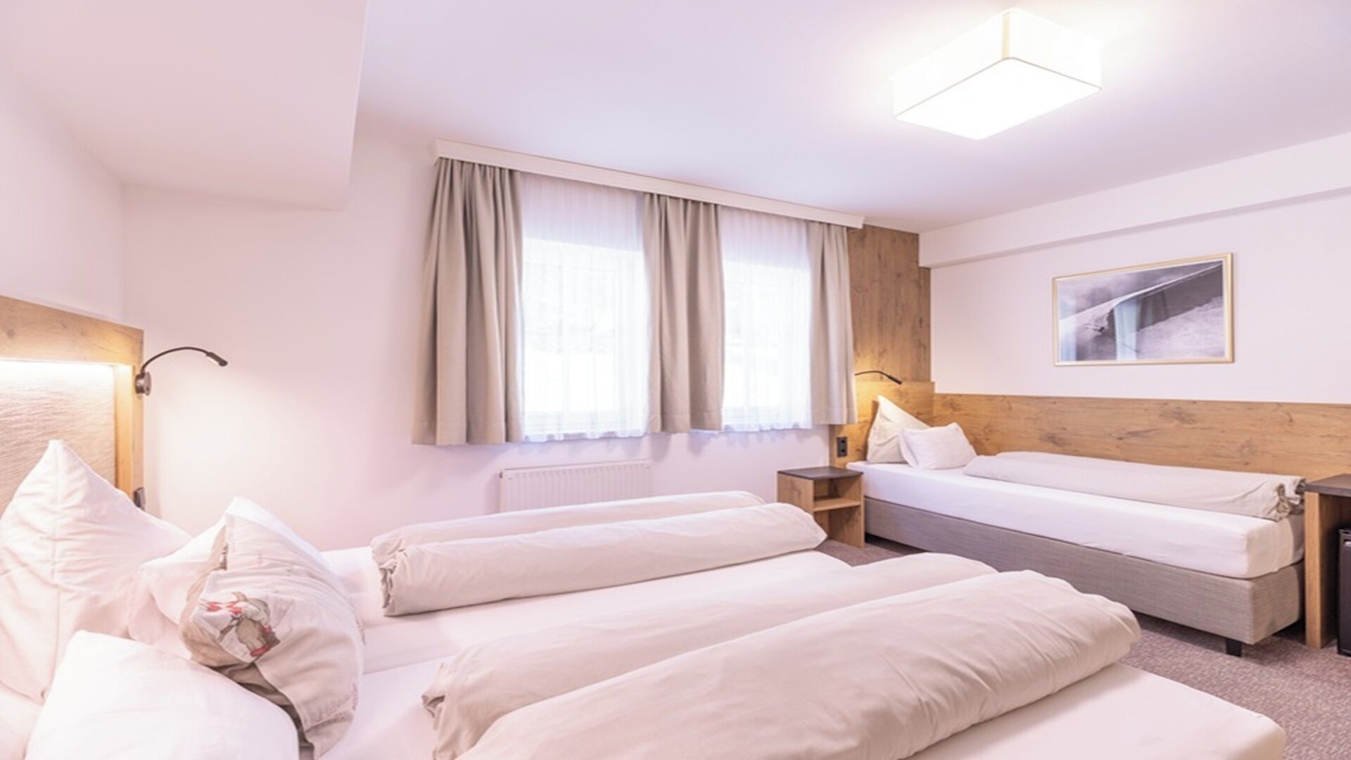 A hotel room with two beds, a wooden headboard, a window with curtains, a bedside table, a lamp, and a framed picture on the wall.