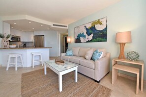 Een moderne keuken met witte kasten en roestvrijstalen apparatuur, een beige bank met blauw-witte kussens, een glazen salontafel en een houten bijzettafeltje met een lamp.