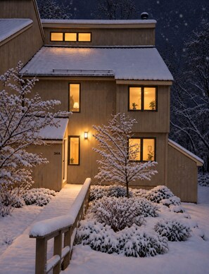 A two-story wooden house with a snow-covered exterior, a visible entrance, and a small porch area.