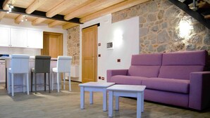 A room with a stone wall, a purple sofa, white tables, and a kitchen area with white cabinets and bar stools. - B&B Tito Livio (Teolo)