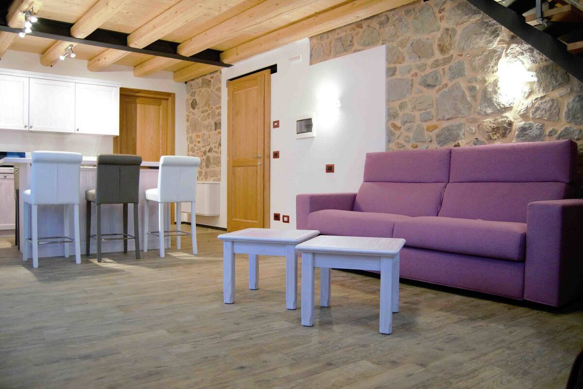 A room with a stone wall, a purple sofa, white tables, and a kitchen area with white cabinets and bar stools.