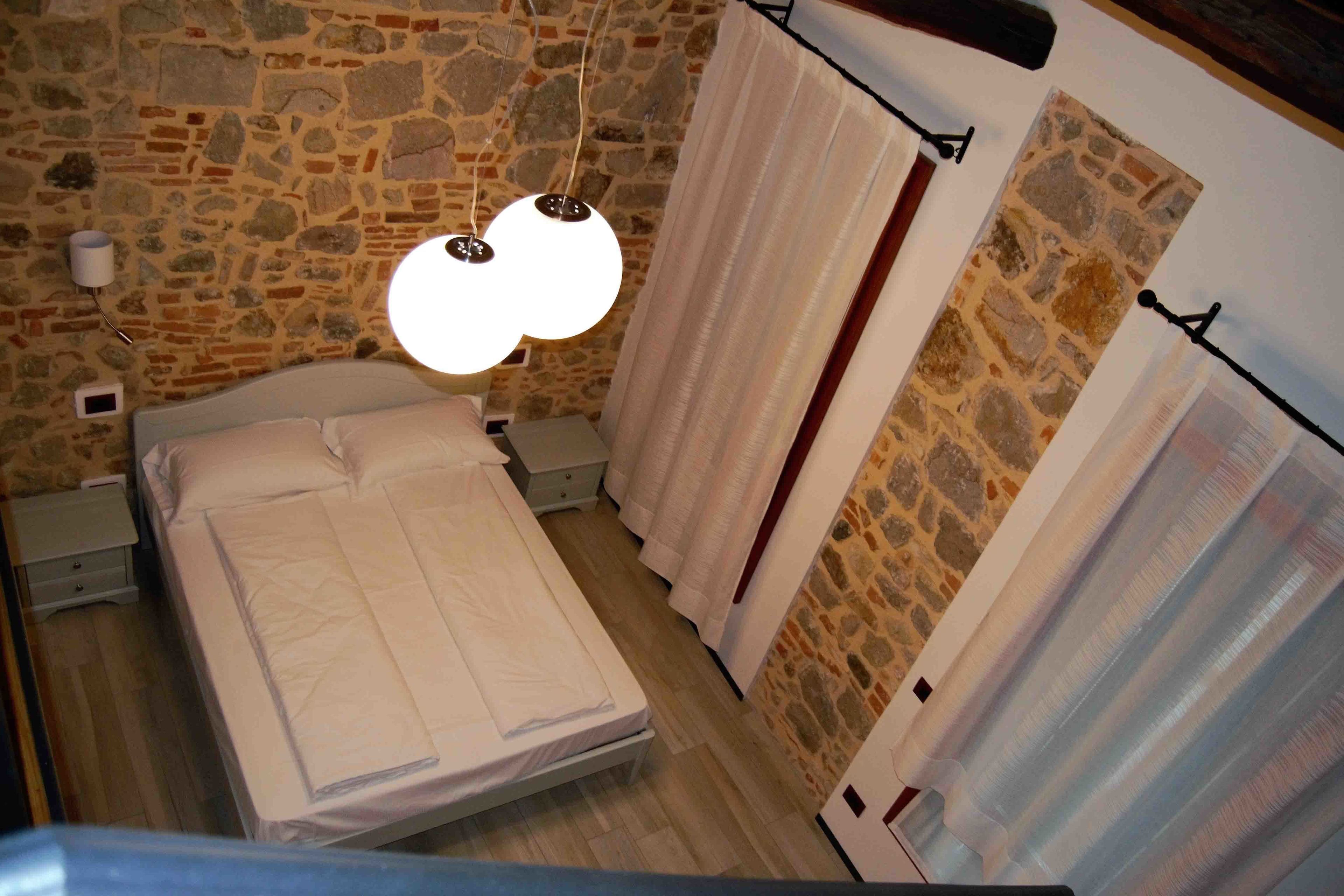 Une chambre avec un mur en pierre, un lit recouvert de draps blancs, un luminaire suspendu et des rideaux.