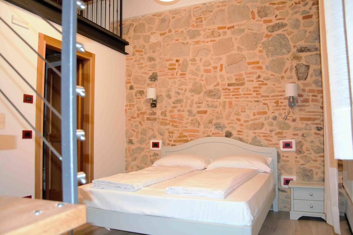 A bedroom with a stone wall, a bed with white linens, two bedside tables, and a wooden door.