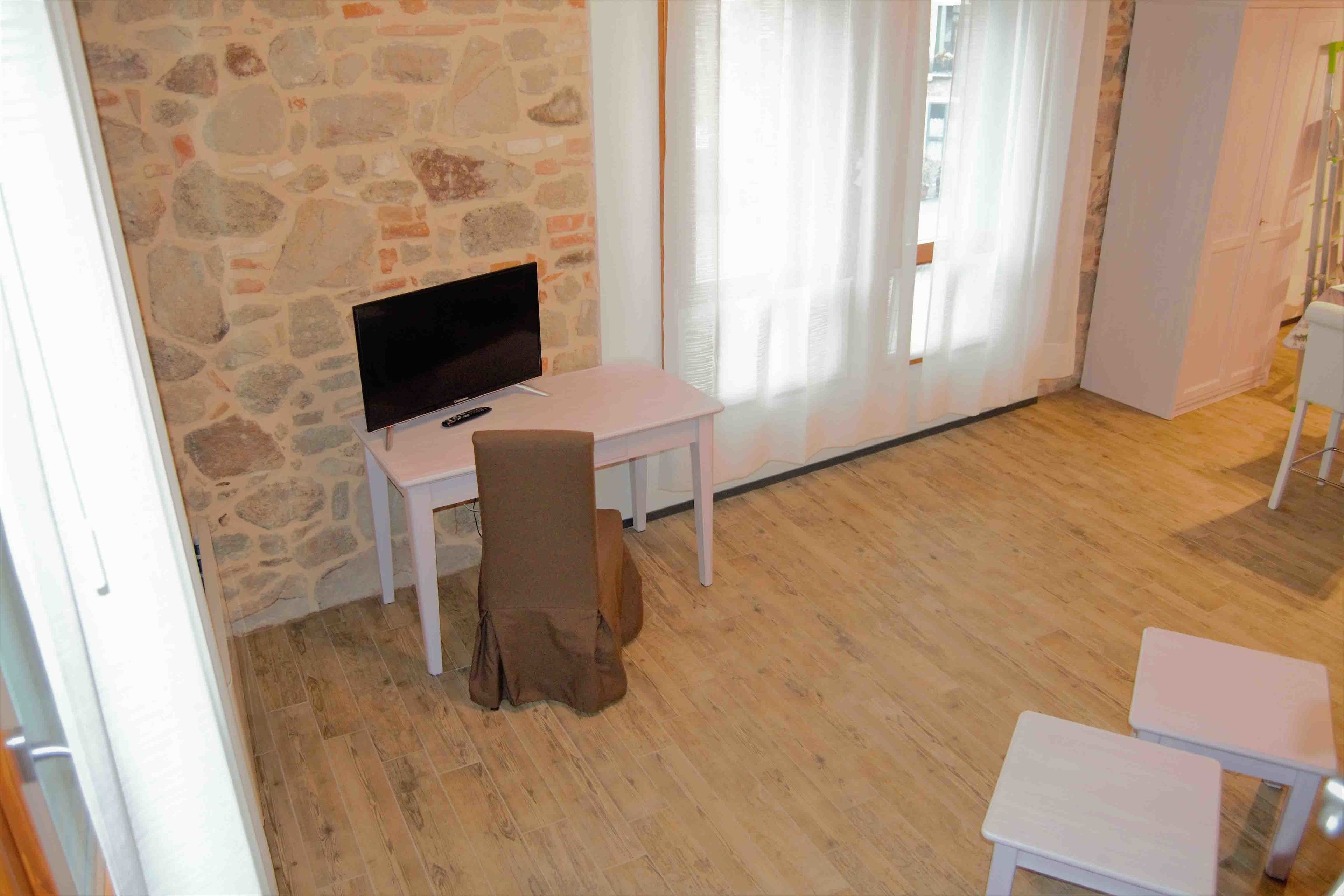 Una habitación con pared de piedra, un escritorio blanco con televisor, una silla marrón y un banco blanco.