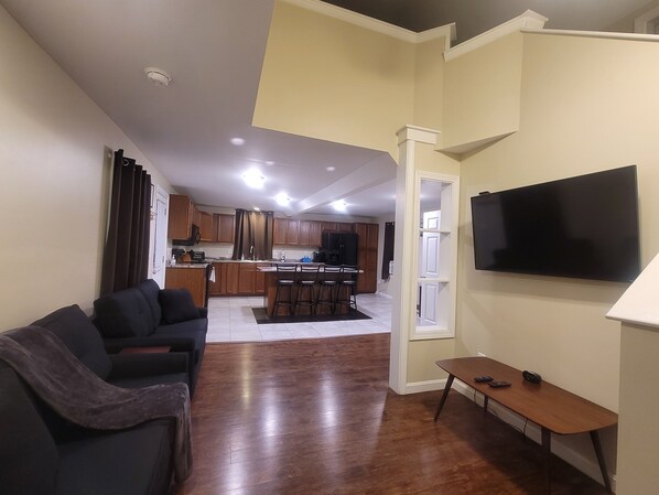 A living room with a sofa, a TV, and a small table. There is a kitchen in the background with wooden cabinets and a dining area.