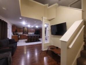 A modern living room with a dark-colored sofa, a wooden dining table with bar stools, a kitchen area with wooden cabinets, and a flat-screen TV mounted on the wall.