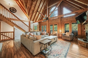 A spacious living room with a wooden staircase, a large sofa, a coffee table, and a television. - Huckleberry Hideout by SoCharm | Private Cabin Oasis (Mooresville)