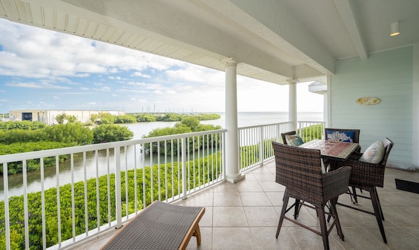 A covered balcony with wicker furniture, a view of a river, and a distant shoreline.