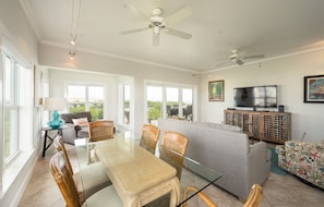 A spacious living room with a glass-top dining table, wicker chairs, a grey sofa, and a television on a wooden cabinet.