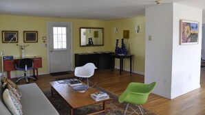 A living room with a sofa, a coffee table, a desk, and a chair. There is a door, a mirror, and a painting on the wall.