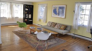 A living room with a sofa, two chairs, a coffee table, a bookshelf, and a large window with curtains.