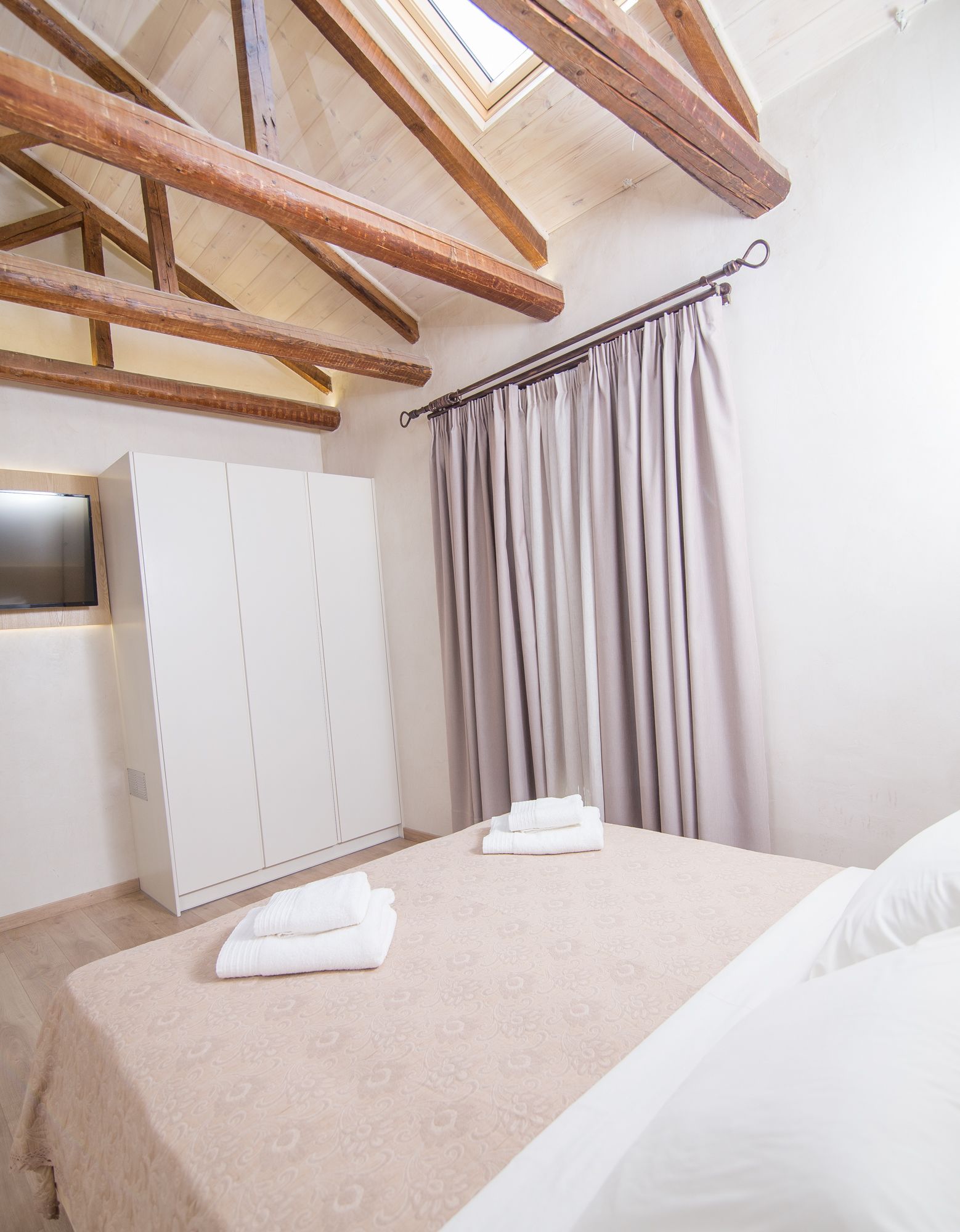 A bedroom with a sloped ceiling, a bed with white linens, a wardrobe, and a window with curtains.