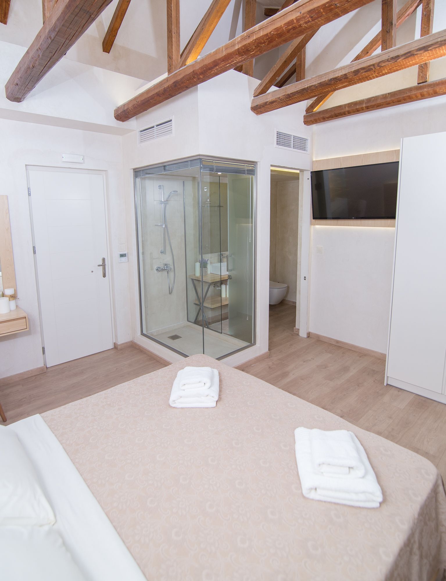 Un baño moderno con cabina de ducha de vidrio, inodoro y lavabo. Hay una cama con colcha color beige y dos toallas dobladas sobre ella.