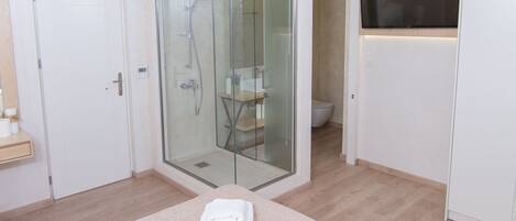 Un baño moderno con cabina de ducha de vidrio, inodoro y lavabo. Hay una cama con colcha color beige y dos toallas dobladas sobre ella.