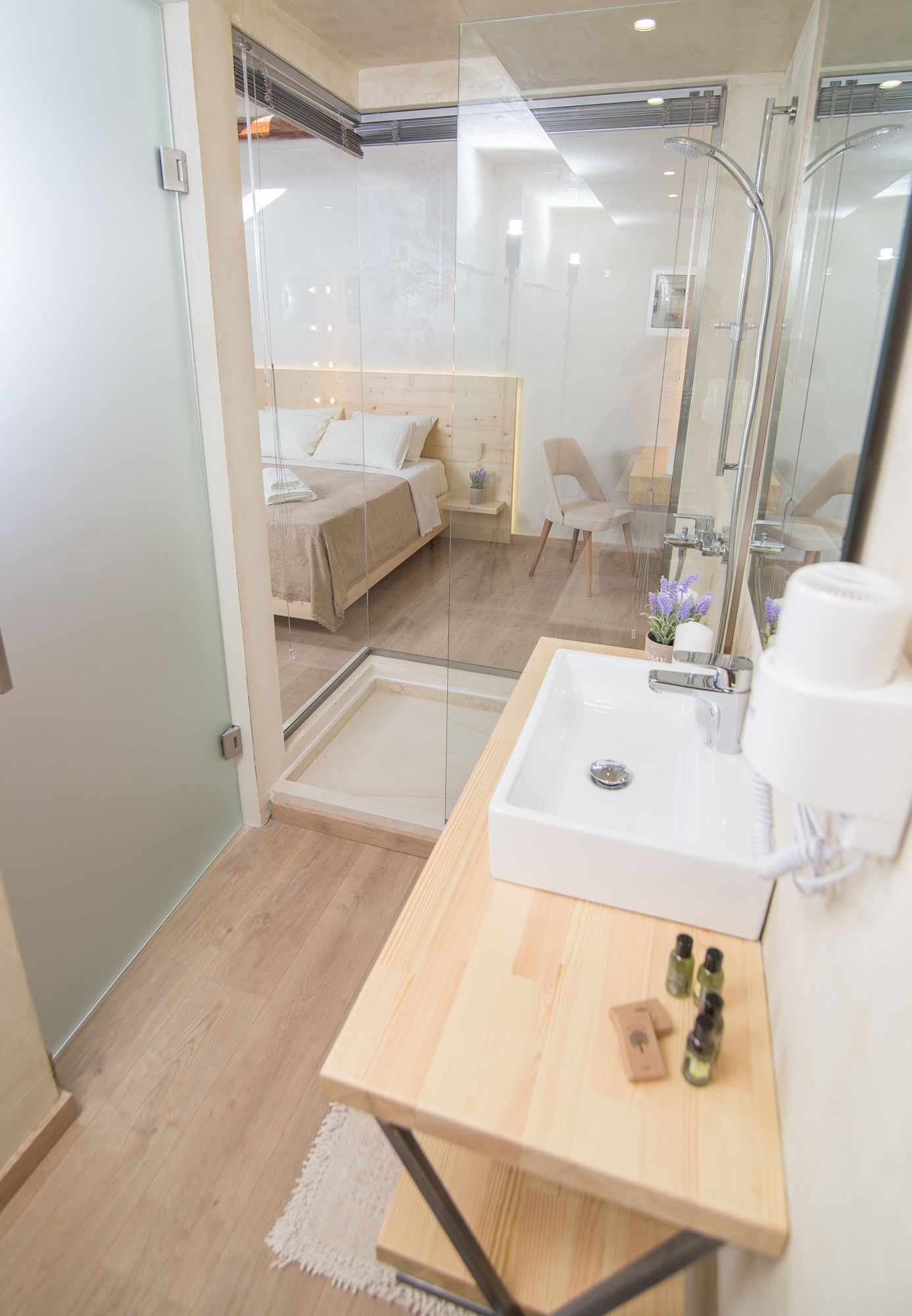 A modern bathroom with a glass shower enclosure, a wooden vanity, and a bed visible through the glass.