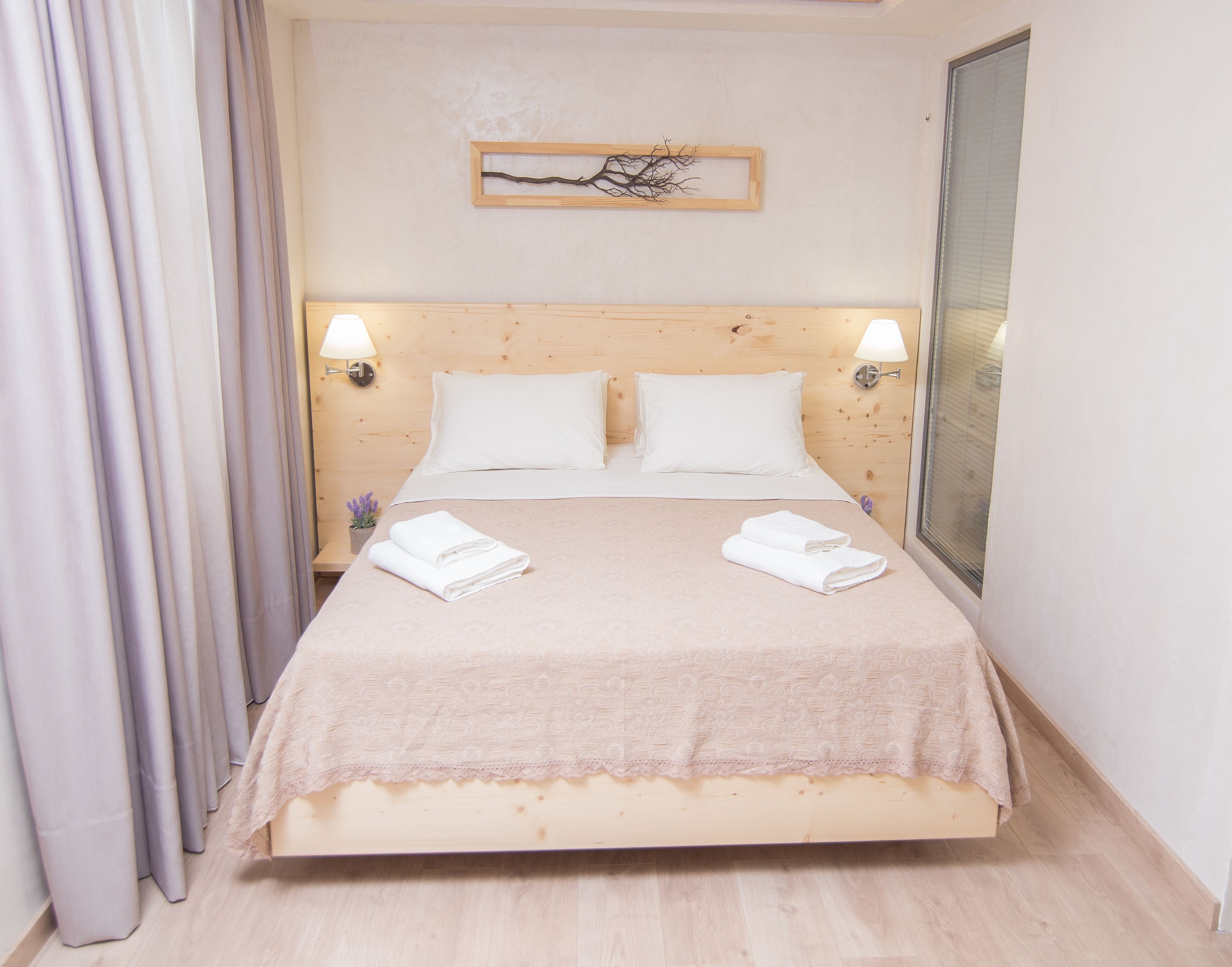 Een netjes opgemaakt bed met witte lakens en twee gevouwen handdoeken, een houten hoofdbord, wandlampen en een ingelijst kunstwerk erboven.